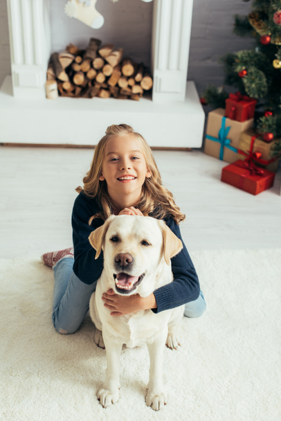happy girl in warm sweater cuddling labrador in decorated living room, christmas concept  - Photo, Image