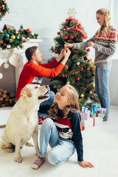 セーターを着た子供の選択的な焦点リビングルームでクリスマスツリーを飾る両親の近くのラブラドールを抱きしめる - 写真・画像