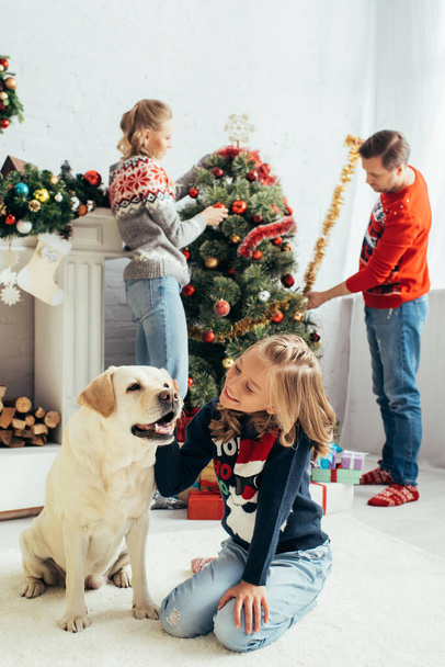 selective focus of joyful girl in sweater cuddling labrador near parents decorating christmas tree in living room - Photo, Image