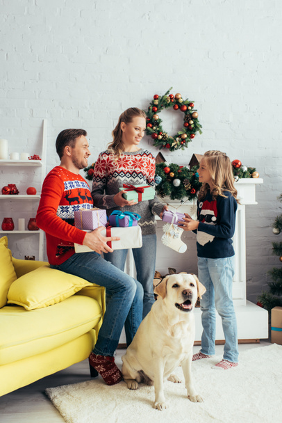 freudige Eltern mit Geschenken an Tochter mit Hund im geschmückten Wohnzimmer  - Foto, Bild