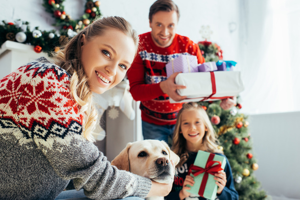 selective focus of happy woman cuddling dog near daughter and husband with gifts on christmas  - Photo, Image