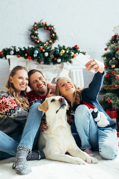 niño tomando selfie con padres alegres y perro en sala de estar decorada  - Foto, imagen