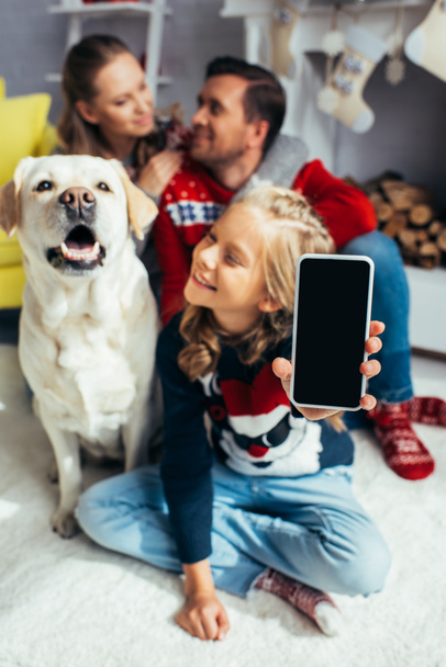 happy girl holding smartphone with blank screen and blurred family on background - Photo, Image