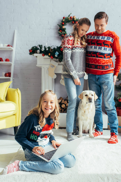 szczęśliwy dzieciak za pomocą laptopa w pobliżu radosnych rodziców stojących w pobliżu psa na Boże Narodzenie - Zdjęcie, obraz