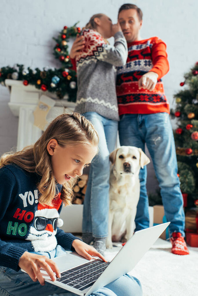 onnellinen lapsi käyttää kannettavaa tietokonetta lähellä vanhempia seisoo lähellä koira ja joulukuusi - Valokuva, kuva