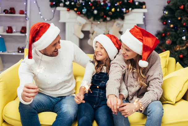 glückliche Familie in Weihnachtsmützen mit Wunderkerzen im geschmückten Wohnzimmer an Weihnachten - Foto, Bild