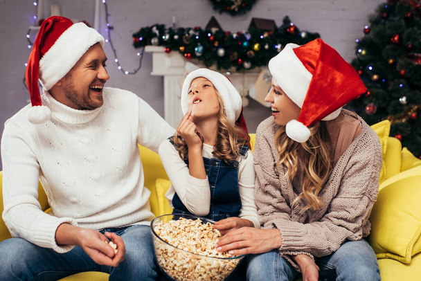Glückliche Eltern in Weihnachtsmannmützen schauen ihrer Tochter beim Popcorn-Essen zu  - Foto, Bild