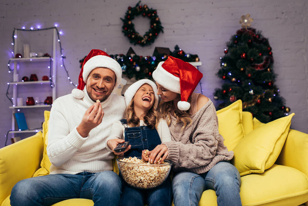 glückliche Mutter und Tochter in Weihnachtsmannmützen lachen neben Mann, der Popcorn isst - Foto, Bild