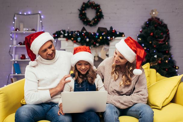 fröhliches Kind mit Weihnachtsmütze und Laptop in der Nähe der Eltern im dekorierten Wohnzimmer  - Foto, Bild