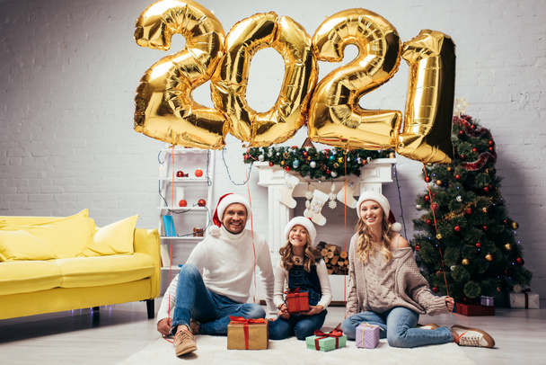 joyful family in santa hats sitting near presents and shiny balloons with 2021 numbers  - Foto, afbeelding