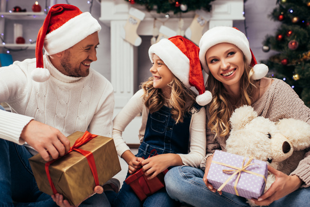 familia complacida en sombreros de santa celebración de regalos en la sala de estar decorada en Navidad  - Foto, Imagen