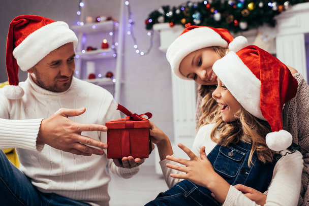 father in santa hat giving present to happy daughter near wife - Фото, зображення