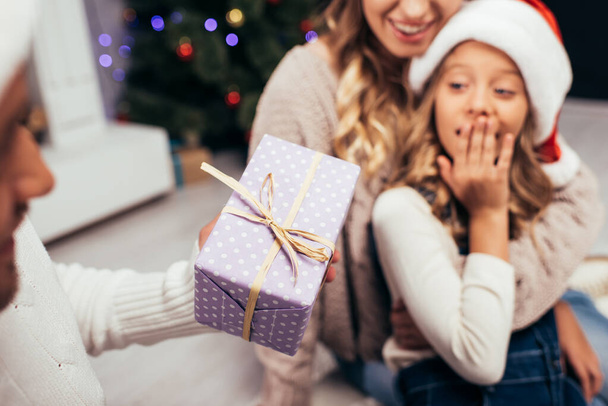 happy father in santa hat giving present to surprised daughter with blurred wife on background - Foto, Bild