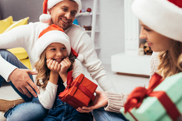 Noel Baba şapkalı anne, babasının yanındaki mutlu kızına hediye veriyor. - Fotoğraf, Görsel