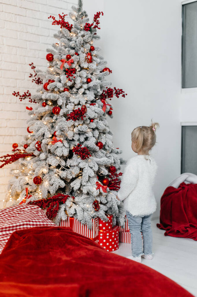 kleines Mädchen im weißen Pullover neben einem Weihnachtsbaum in roten Farben - Foto, Bild