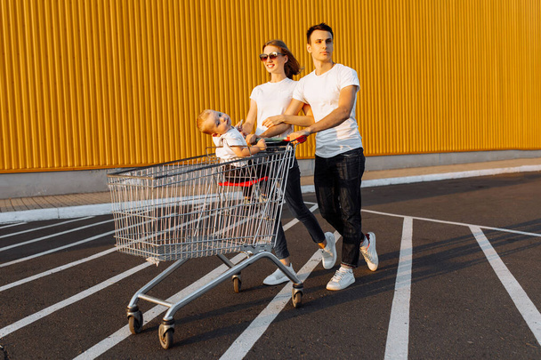 Happy family, man and woman having fun while rolling their baby in shopping trolley, outdoors, shopping concept - Photo, image