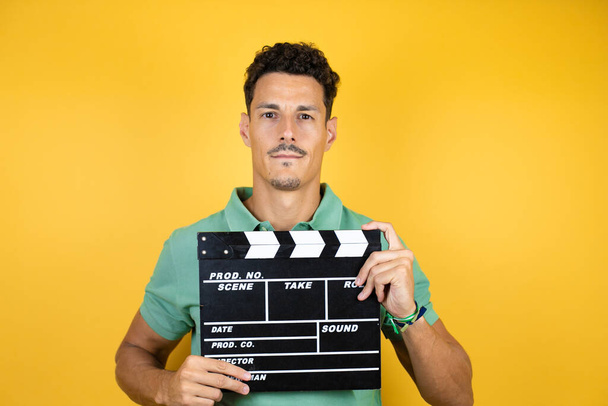 Young handsome man wearing green casual t-shirt over isolated yellow background holding clapperboard very happy - Photo, Image