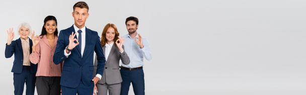 Positive multiethnic businesspeople looking at camera while showing ok gesture isolated on grey, banner  - Фото, изображение