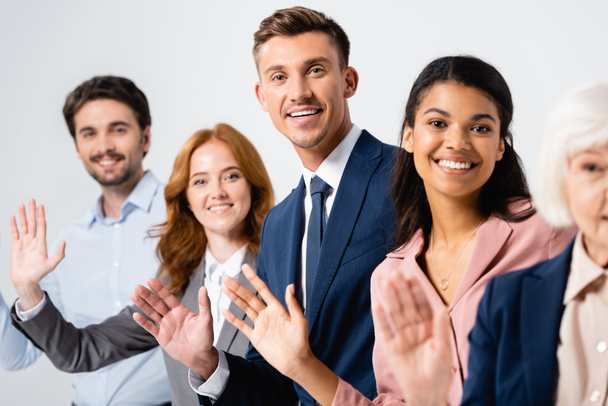 Veselý podnikatel mává rukou poblíž multikulturních kolegů na rozmazaném pozadí izolovaném na šedém - Fotografie, Obrázek