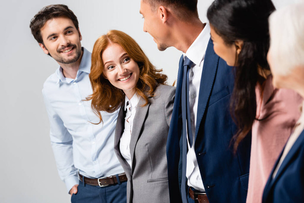Mujer de negocios sonriente mirando a colegas multiétnicos en primer plano borroso aislado en gris - Foto, Imagen