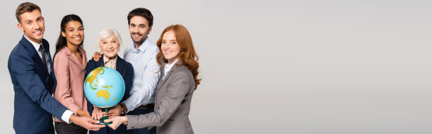 Smiling multiethnic businesspeople holding globe and looking at camera isolated on grey, banner  - Photo, Image