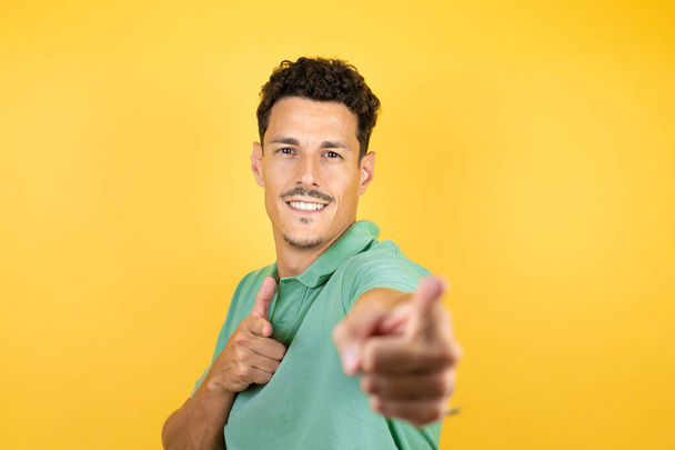 Mladý pohledný muž v zeleném ležérním tričku na izolovaném žlutém pozadí ukazuje prstem na vás a kameru, usmívá se pozitivně a vesele - Fotografie, Obrázek