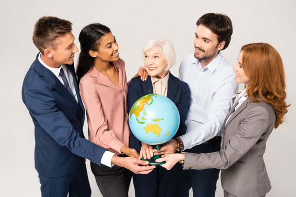 Multicultural businesspeople smiling and embracing while holding globe isolated on grey - Photo, Image