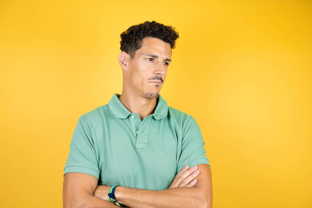 Junger gutaussehender Mann in grünem lässigem T-Shirt vor isoliertem gelben Hintergrund skeptisch und nervös, missbilligender Gesichtsausdruck mit verschränkten Armen - Foto, Bild