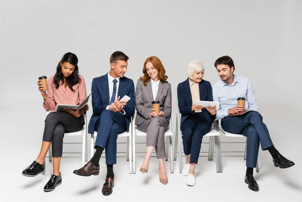 Multiethnic businesspeople with coffee to go using devices on chairs on grey background - Fotografie, Obrázek