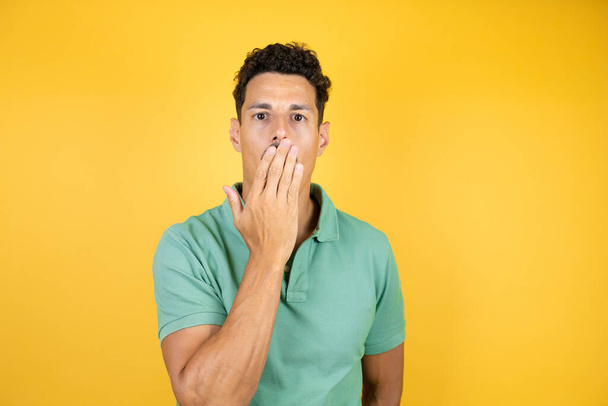 Joven hombre guapo vistiendo camiseta casual verde sobre fondo amarillo aislado sorprendido cubriendo su boca con su mano - Foto, imagen