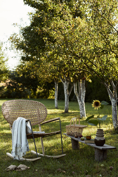 Stylish composition of countryside garden with design rattan armchair, wooden bench, plaid, food, drinks and elegant accessories. A lot of colorful flowers. Summer mood.  - Фото, изображение