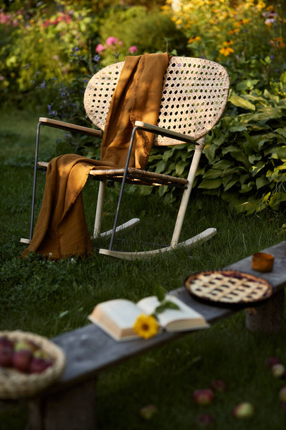 Stylish composition of countryside garden with design rattan armchair, wooden bench, plaid, food, drinks and elegant accessories. A lot of colorful flowers. Summer mood.  - Photo, image