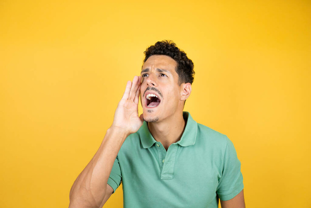 Mladý pohledný muž v zeleném neformálním tričku přes izolované žluté pozadí křičel a křičel hlasitě na stranu s rukou na ústech. Koncept komunikace. - Fotografie, Obrázek