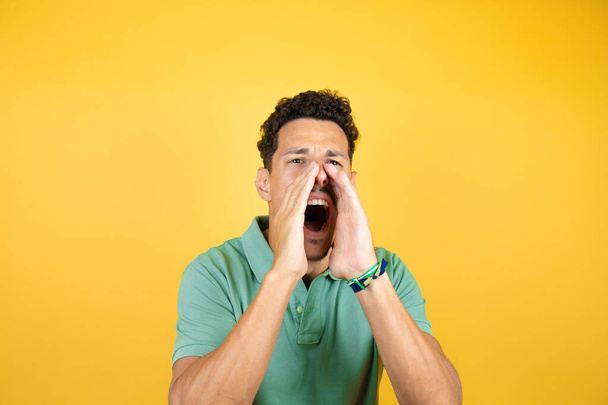 Jonge knappe man draagt groen casual t-shirt over geïsoleerde gele achtergrond schreeuwen en schreeuwen luid naar zij met handen op de mond. Communicatieconcept. - Foto, afbeelding