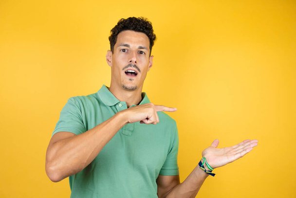 Jonge knappe man draagt groen casual t-shirt over geïsoleerde gele achtergrond verbaasd kijken naar de camera presenteren iets op de hand en wijzen met de vinger. - Foto, afbeelding