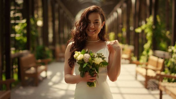 Cheerful bride posing with bouquet under arch. Lady waiting for wedding ceremony - Фото, изображение