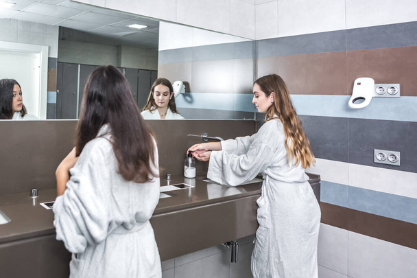 Takaisin näkymä nuori nainen kylpytakki pestä kädet yli pesuallas lähellä tyttöystävä kampaus hiukset kylpyhuoneessa moderni kylpylä salon - Valokuva, kuva