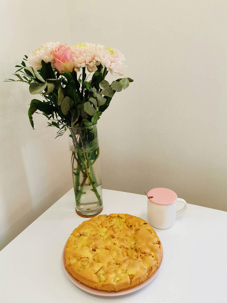 Gemütliche Komposition in der Küche. Köstliche Charlotte, ein Strauß Rosen und Chrysanthemen auf einem weißen Tisch. Tischdekoration mit einem Blumenstrauß zum Frühstück und Mittagessen. Hausgemachtes Backkonzept. - Foto, Bild