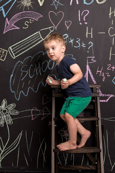 Ein dreijähriger Junge zeichnet mit mehrfarbiger Kreide auf eine große Tafel. schwarze Wand mit Kinderzeichnungen und Formeln. Vertikale Fotografie. - Foto, Bild