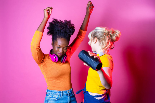 若い美しい黒と白亜の女性がピンクのバッグラウンドにワイヤレススピーカーを保持して屋内で踊る- 2つの孤立した多様な女性の友人クラブ-幸福、興奮、ダンスコンセプト - 写真・画像