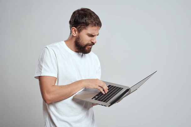 Een man met een laptop in zijn handen op een lichte achtergrond in een wit t-shirt emoties licht achtergrond nieuwe technologieën - Foto, afbeelding