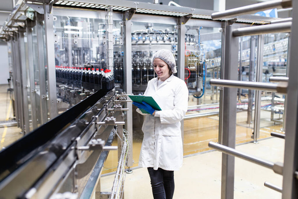 Jonge vrolijke vrouwelijke werknemer in de bottelfabriek controleert sapflessen voor verzending. Inspectiekwaliteitscontrole.  - Foto, afbeelding