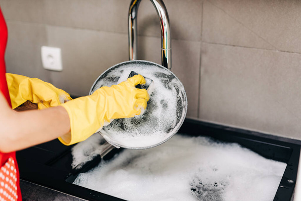 Primo piano della mano della donna in guanti di gomma protettiva gialla lavare i piatti in cucina. - Foto, immagini