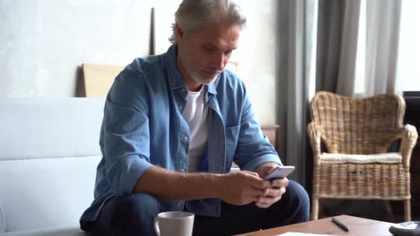 Middle aged man with grey hair sitting on the sofa using phone, texting message - Materiaali, video