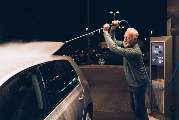 Senior uomo lavare la sua auto la sera alla stazione di lavaggio auto con acqua ad alta pressione. - Foto, immagini