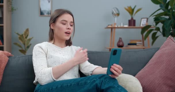 Pretty woman showing with with deaf-mute sign language phrase I miss you while sitting on sofa.Millennial female person using smartphone while having video call. Hearing loss. - Materiał filmowy, wideo