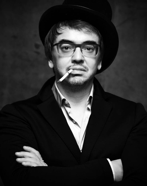 Portrait of middle-aged unshaved man in glasses hipster in black jacket, white shirt and stylish hat smoking cigarette - Fotografie, Obrázek