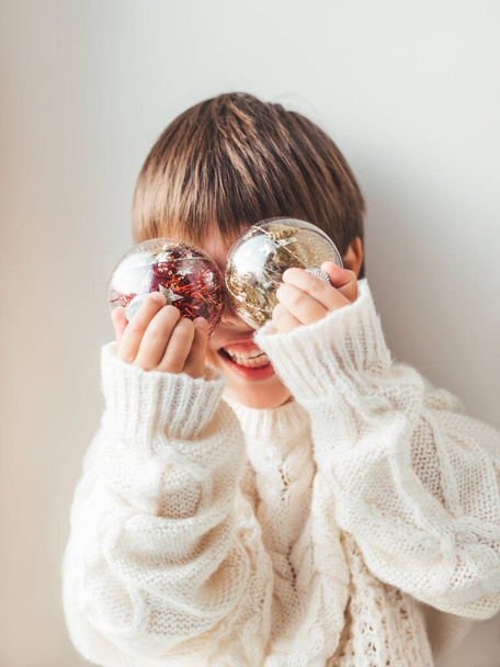 Kid with decorative balls for Christmas tree.Boy in cable-knit oversized sweater.Cozy outfit for snuggle weather.Transparent balls with red, golden spangles inside.Winter holiday spirit.New year. - Foto, Bild