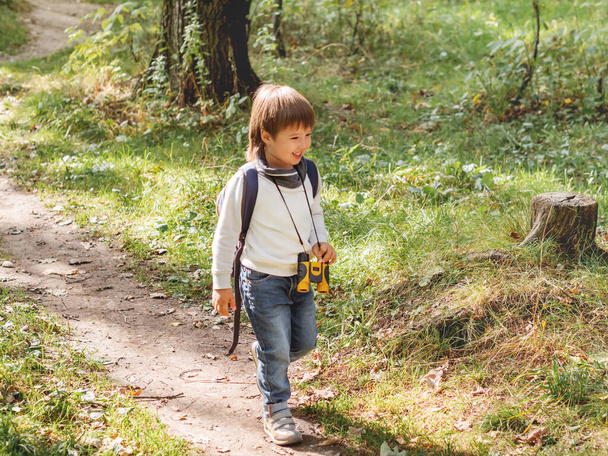 Curious boy is hiking in forest lit by sunlight. Outdoor leisure activity for kids. Child with binoculars and backpack. - Zdjęcie, obraz