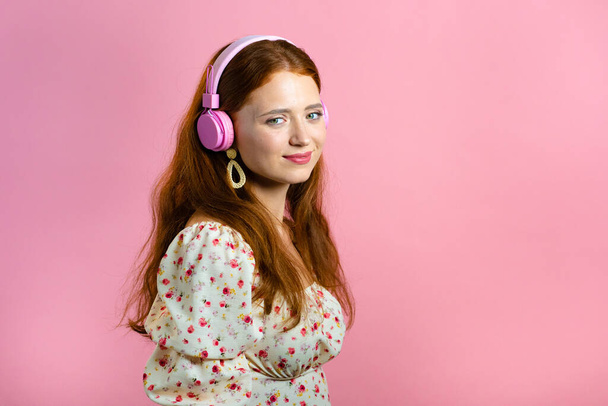 Attractive woman with red hair with headphones on pink studio background. Cute girl portrait. Music, radio, happiness, freedom, youth concept. - Zdjęcie, obraz